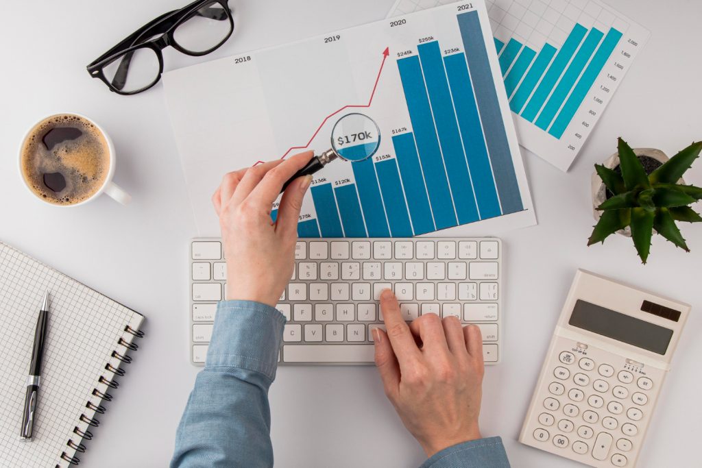 Person analyzing a financial chart with a magnifying glass on a desk with a cup of coffee, calculator, glasses, and notepad.